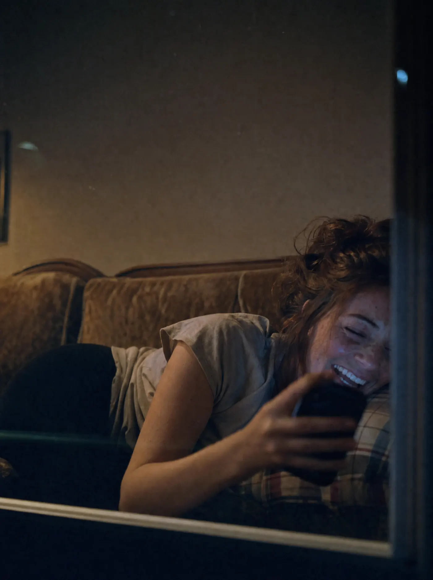 Through a window at night, a woman on a sofa laughs at her phone.